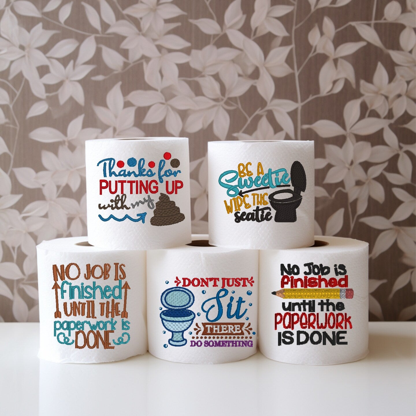 A stack of white toilet paper rolls with various humorous embroidered messages stitched on them, placed against a leafy background.