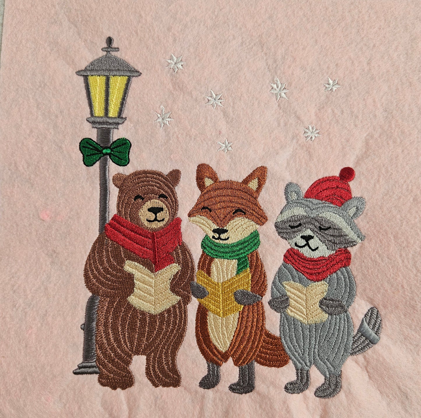 Embroidered design of three animals (bear, fox, and racoon) singing carols next to a streetlamp on a textured beige background.