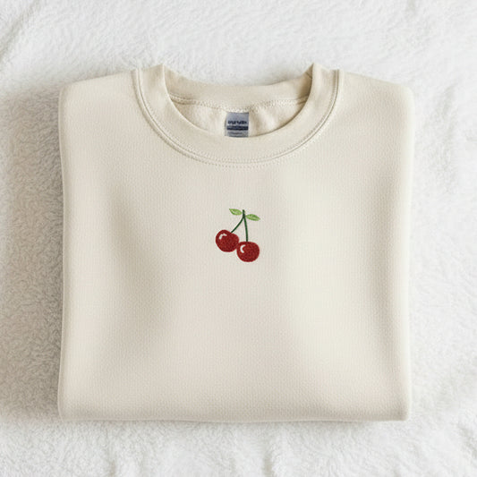 Folded sand-colored sweatshirt with cherry design on a textured white surface