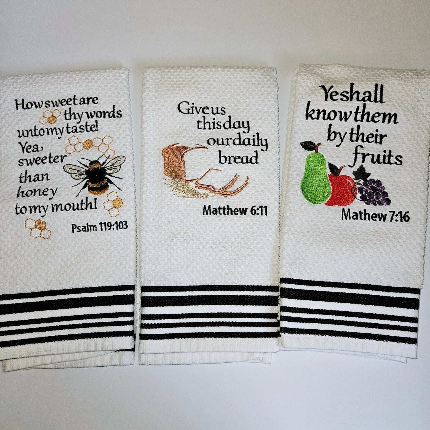 Three white kitchen towels with embroidered Bible verses and accompanying images, displayed in a row with a striped pattern at the bottom.