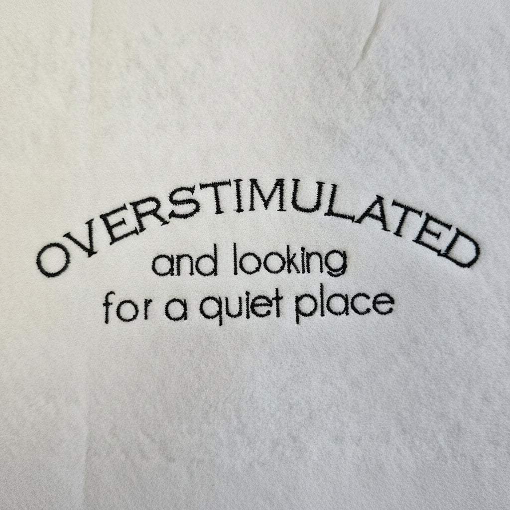 Embroidered text 'OVERSTIMULATED and looking for a quiet place' on a textured white background