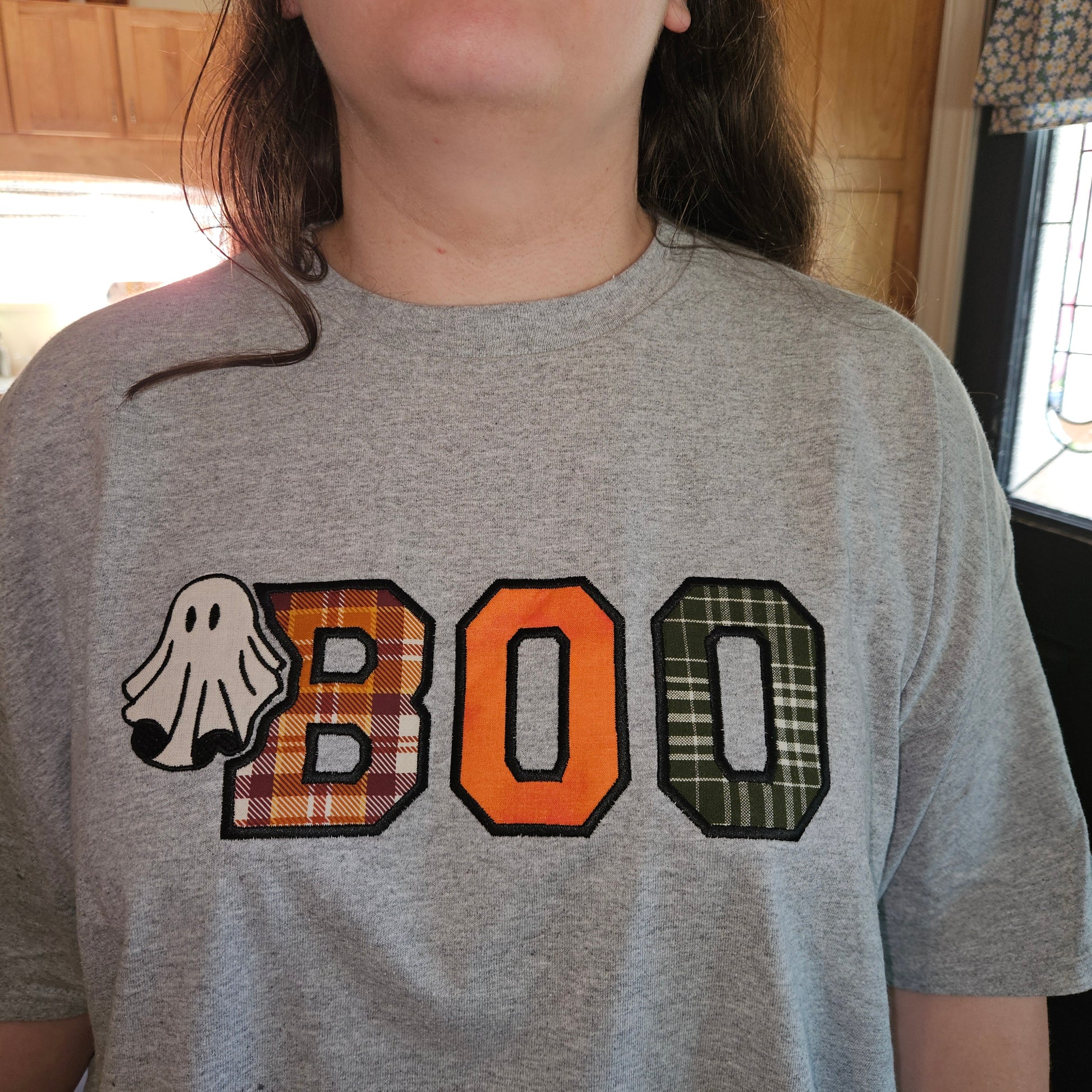 A person wearing a heather gray t-shirt with a 'BOO' graphic. The letter 'B' is filled with a red and brown plaid pattern and a white ghost graphic is on the left.