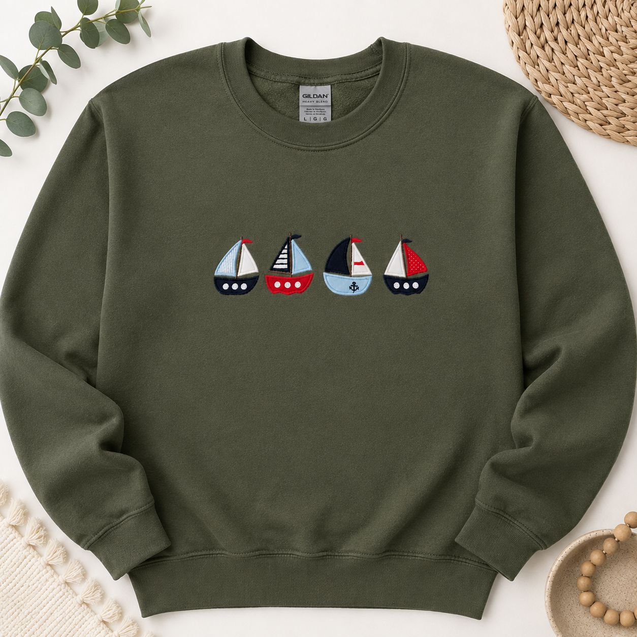 An military green crewneck sweatshirt featuring a row of four colorful sailboats across the chest, styled in a flat-lay arrangement with decorative eucalyptus leaves, a woven hat, and a wooden bead bowl.