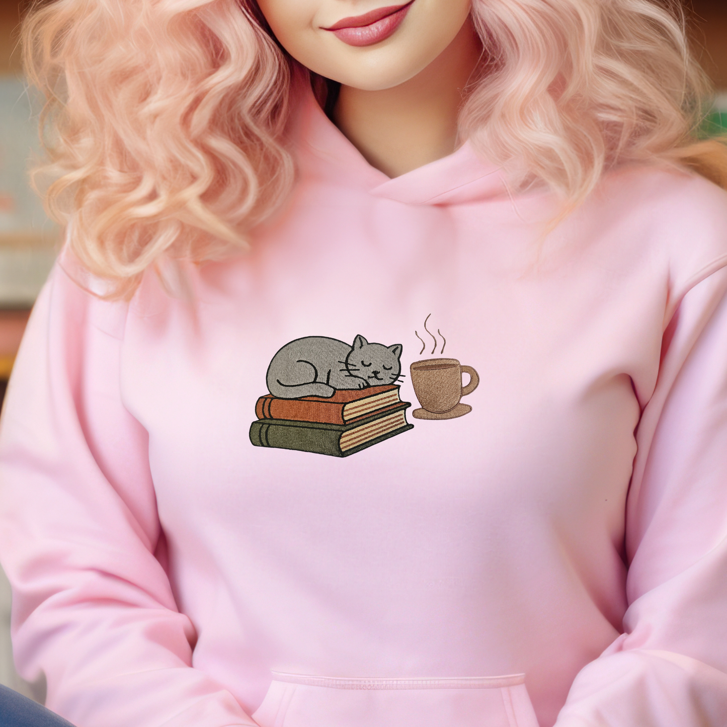 Person wearing a light pink hoodie with a design of a cat, books, and a coffee cup.