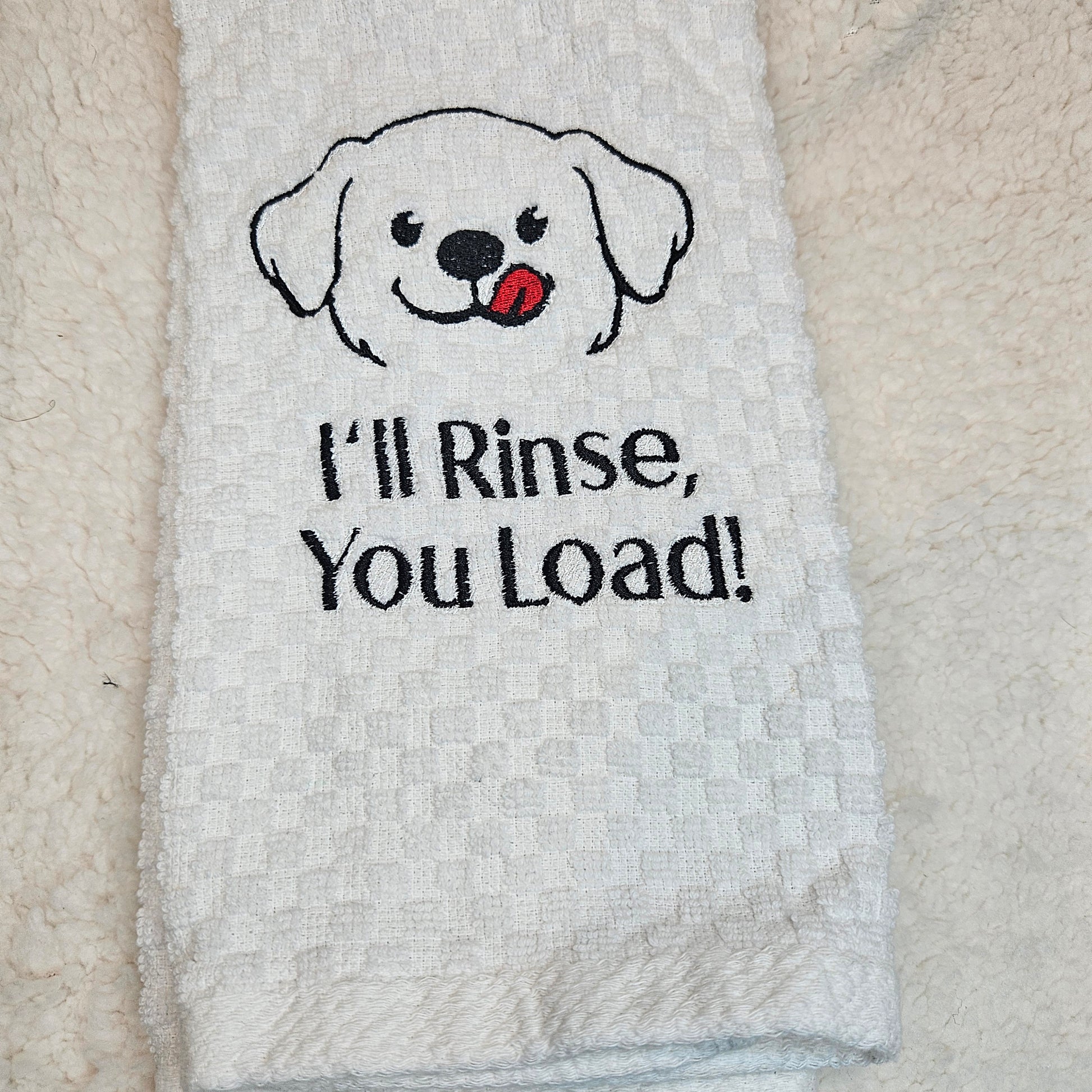 White towel with a dog illustration and text 'I'll Rinse, You Load!' on a textured surface.