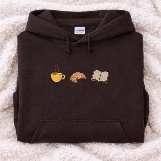 Folded dark chocolate hoodie with coffee cup, croissant, and book design on a textured surface