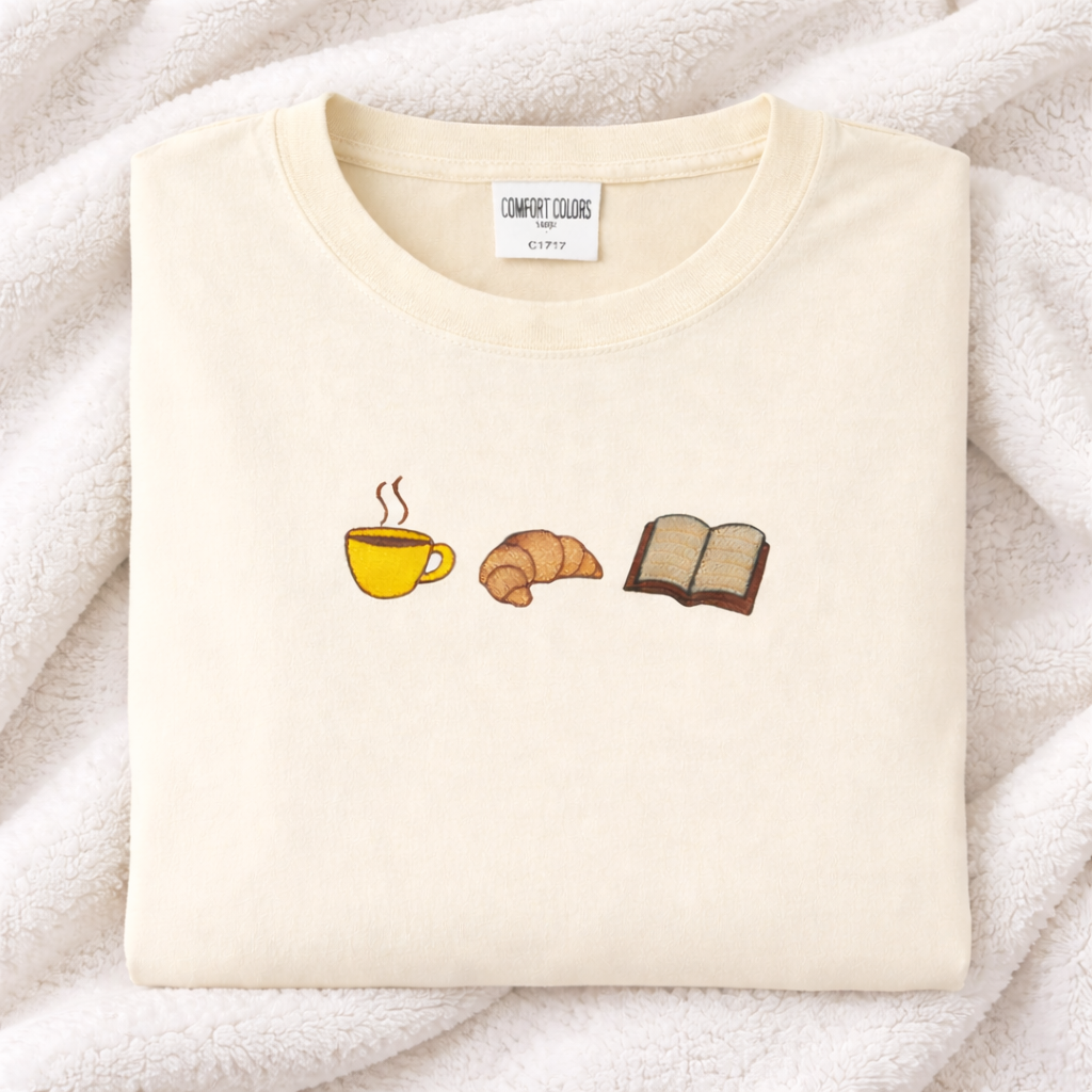 Ivory t-shirt with coffee cup, croissant, and book emoji designs on a soft white background.