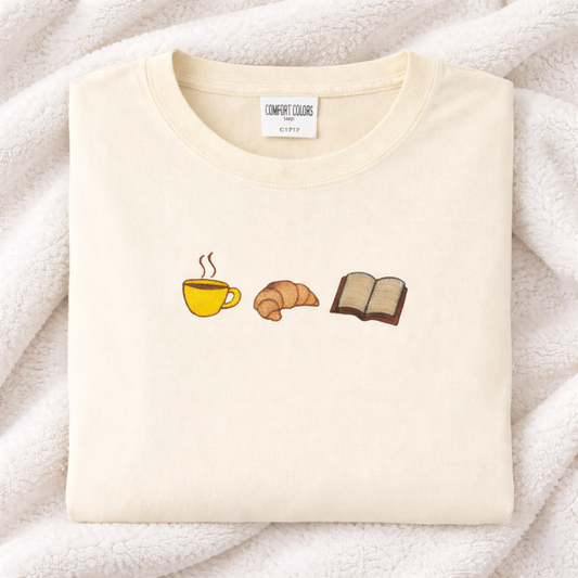 Ivory t-shirt with coffee cup, croissant, and book emoji designs on a soft white background.