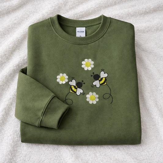 Folded military green sweatshirt with bee and flower designs on a textured surface