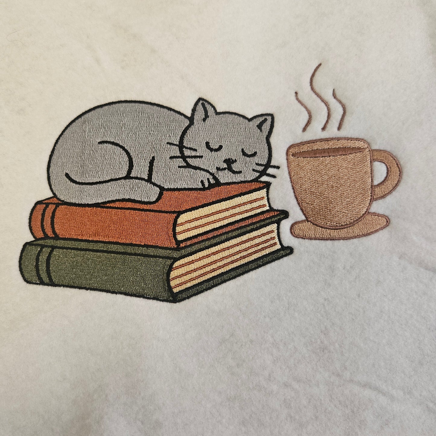 Embroidered design of a cat with books and a mug on a fabric background