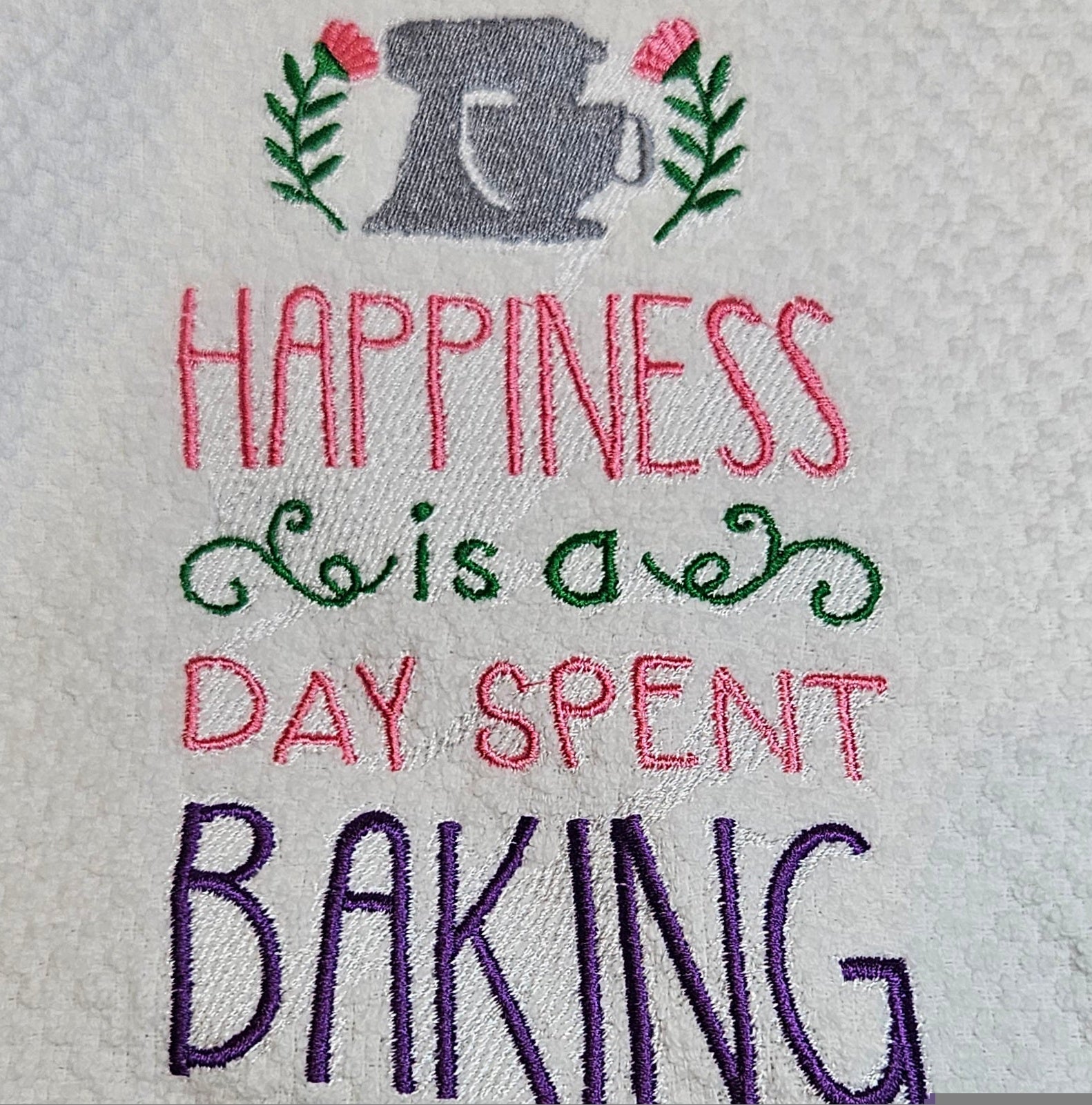 Embroidered text on towel with a mixer design and flowers, reading 'Happiness is a day spent baking'.
