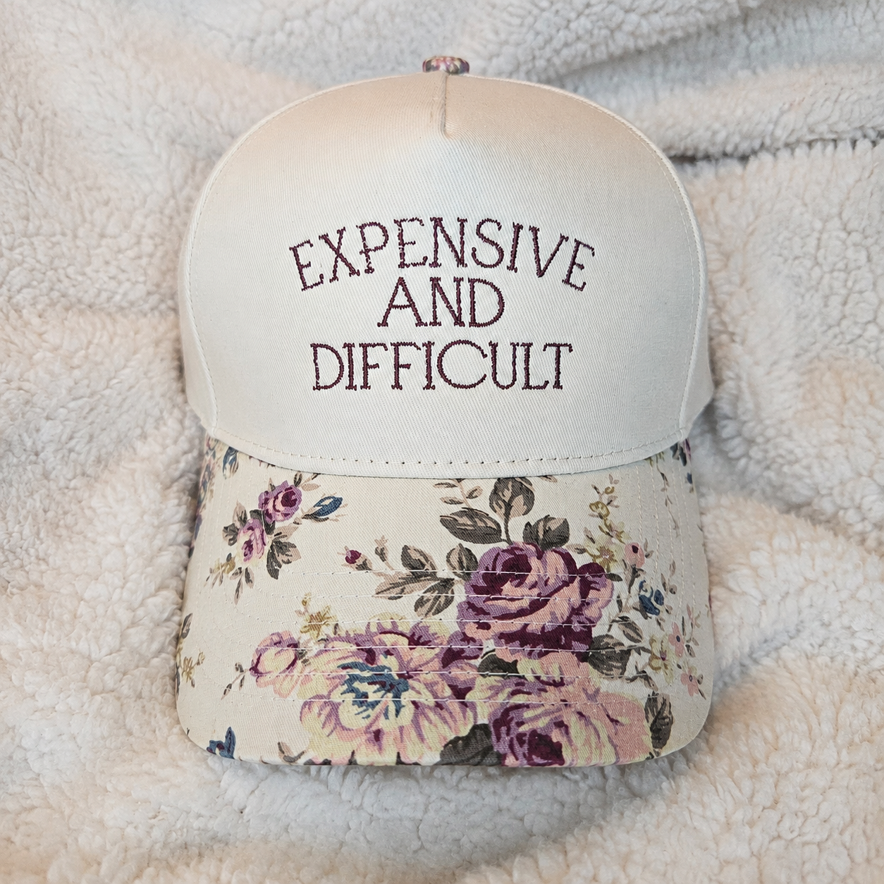 Cap with floral bill and 'EXPENSIVE AND DIFFICULT' text on a soft surface