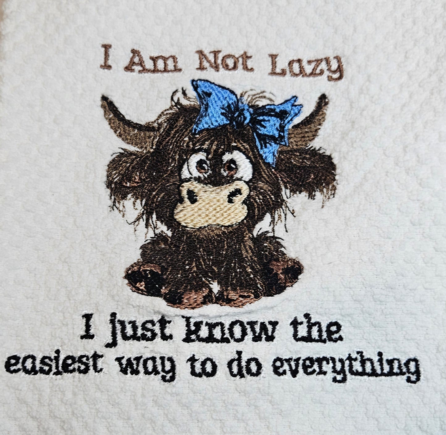 Embroidered design of a cartoon cow with a blue bow and text on a white fabric background.