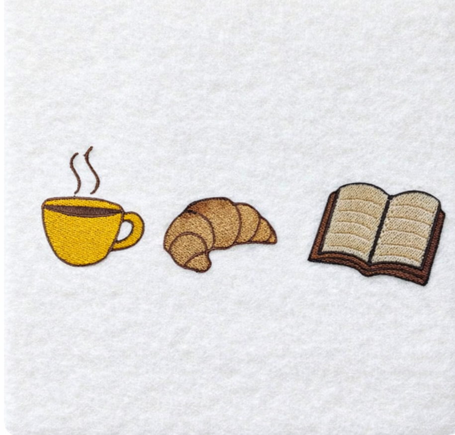 Embroidered design on a white fabric with a coffee cup, croissant, and book.