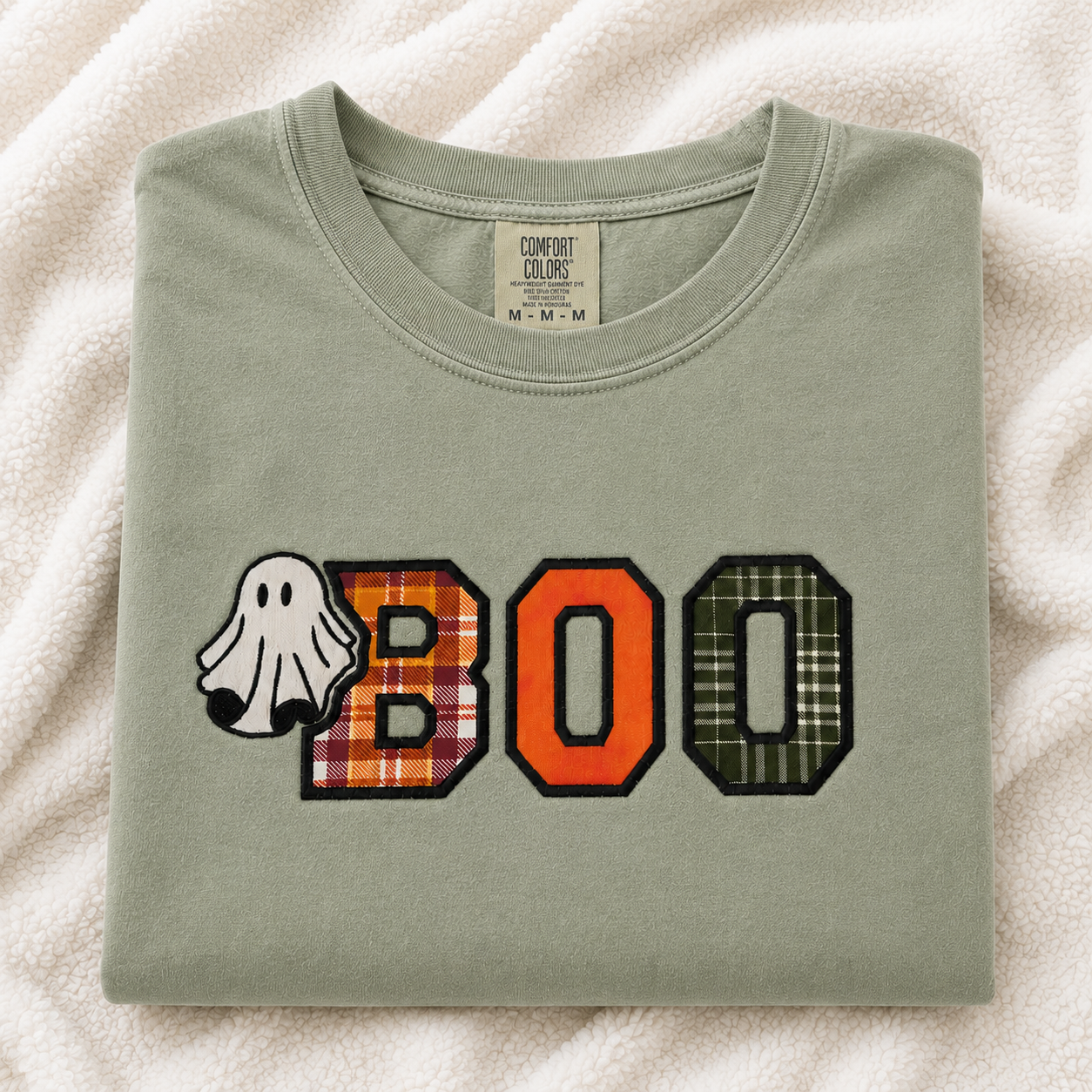 A folded sage green Comfort Colors t-shirt with a 'BOO' graphic on the front, featuring a ghost and plaid patterned letters, laid flat on a white textured blanket.