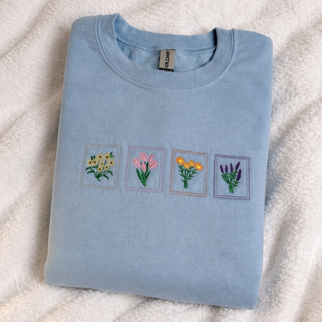 Folded light blue sweatshirt with embroidered flower designs on a textured white surface