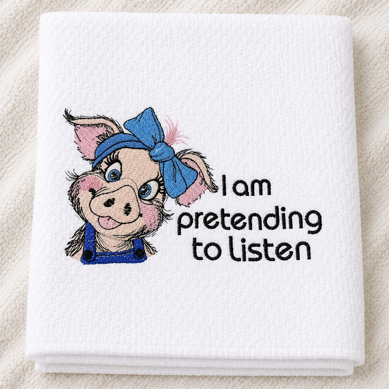 White towel with embroidered pig design and text 'I am pretending to listen' on a beige background