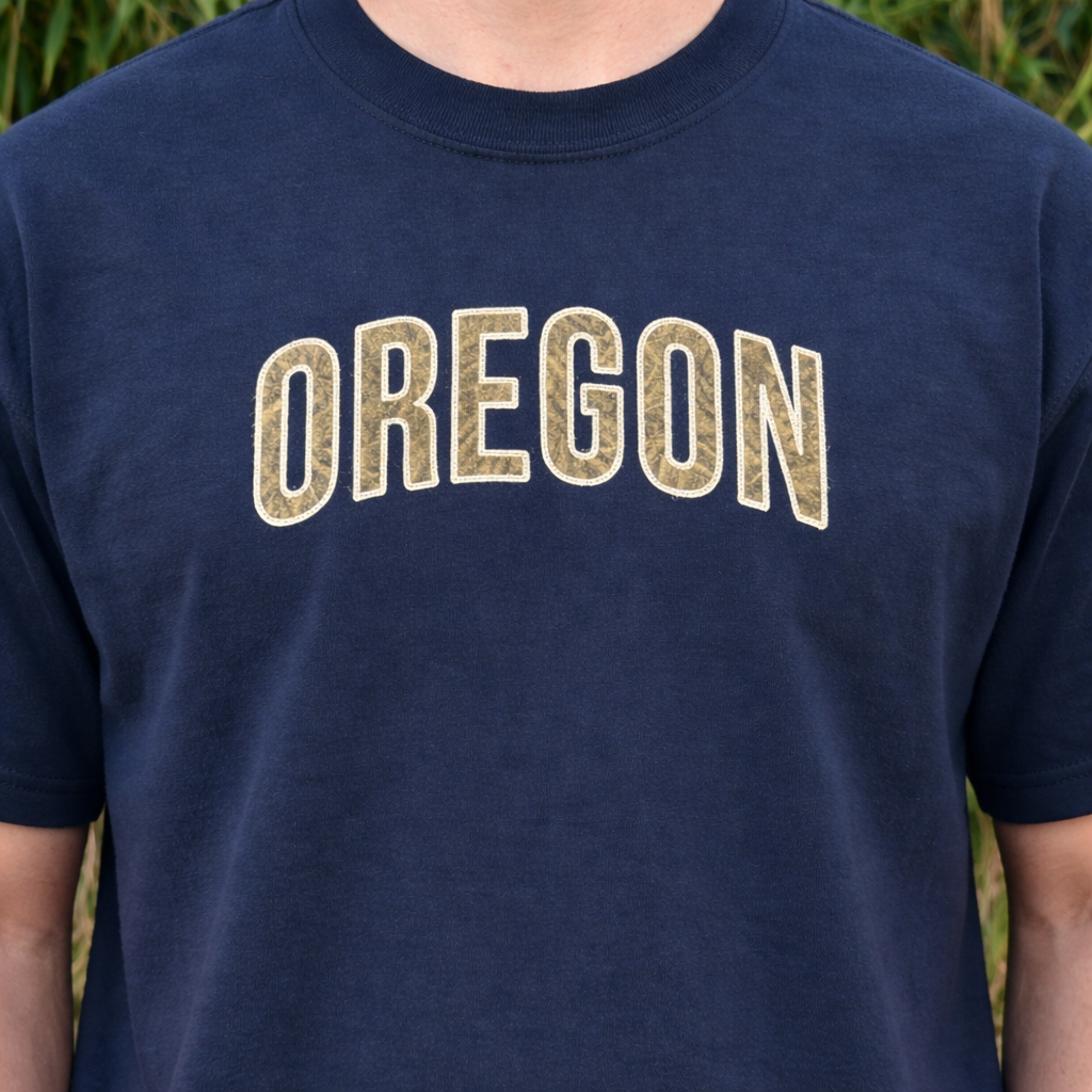 Navy t-shirt with applique 'OREGON' embroidered in beige on a blurred natural background