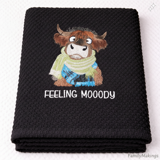 Black towel with a cartoon cow wearing a green and blue scarfs.  With the embroidered text 'Feeling Moody' on a white background.