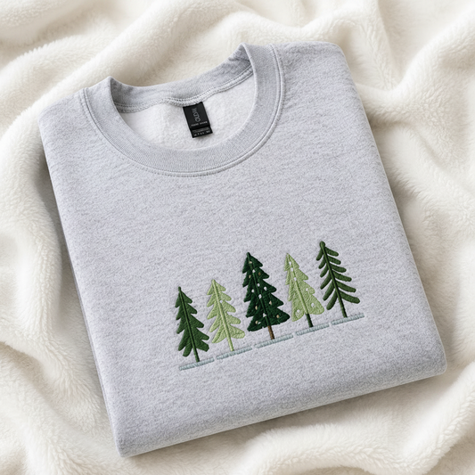 A folded ash gray crewneck sweatshirt with embroidered pine trees on the chest, placed on a soft cream-colored fleece blanket. 