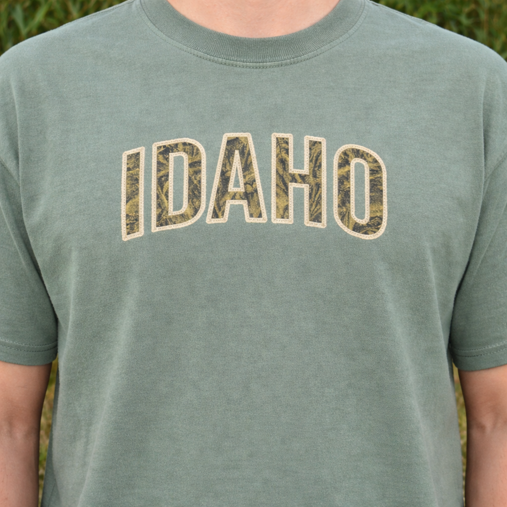 Sage t-shirt with applique 'IDAHO' embroidered on it against a blurred natural background