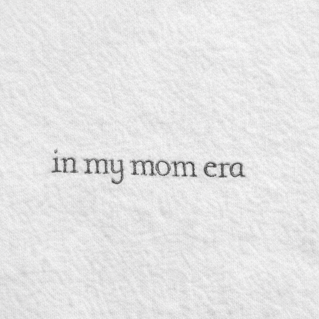Text 'in my mom era' embroidered on a textured white background
