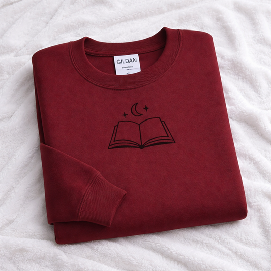 Folded maroon sweatshirt with a black icon of an open book and crescent moon on a white surface