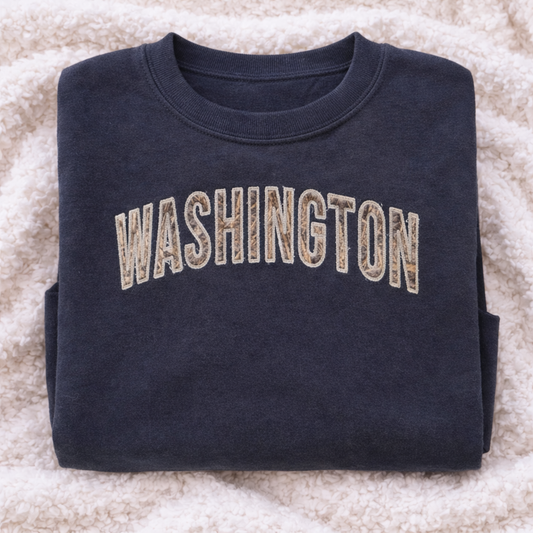 Folded navy sweatshirt with 'WASHINGTON' stitched on it, lying on a textured surface.