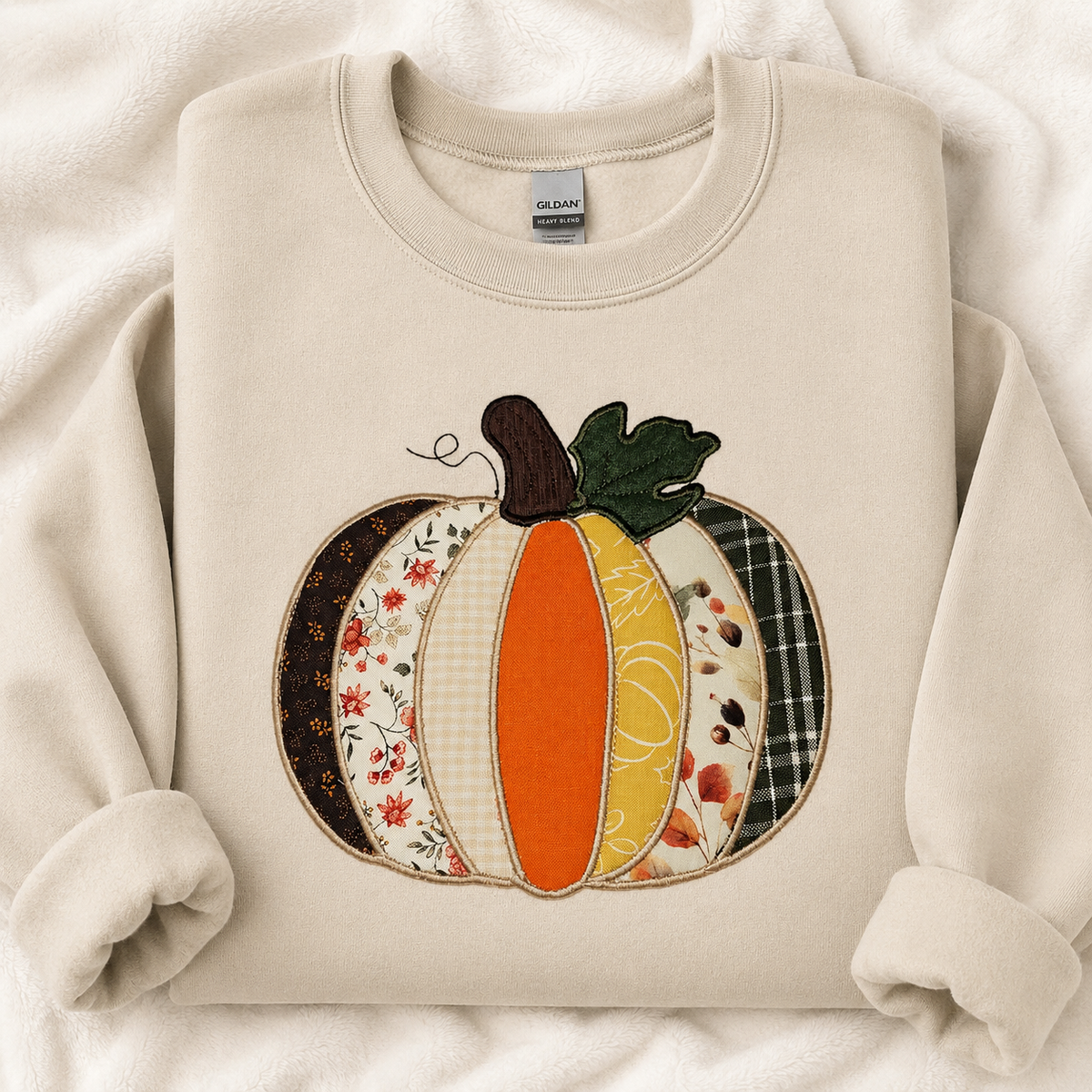 A sand crewneck sweatshirt featuring a decorative applique pumpkin design with autumn-themed patterned panels, laid flat on a soft white textured blanket. The neck label shows the brand 'GILDAN'.