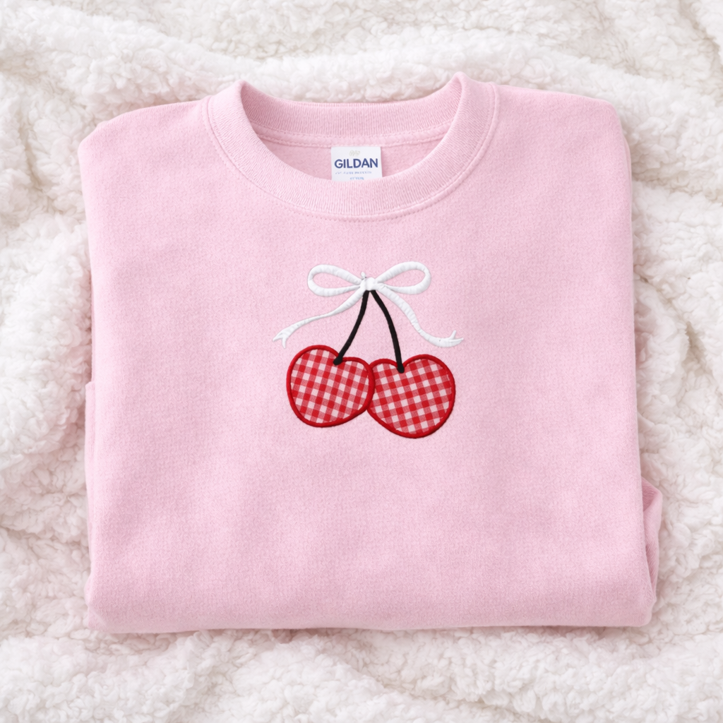 Light pink sweatshirt with red gingham cherries and a white bow on a fluffy white surface