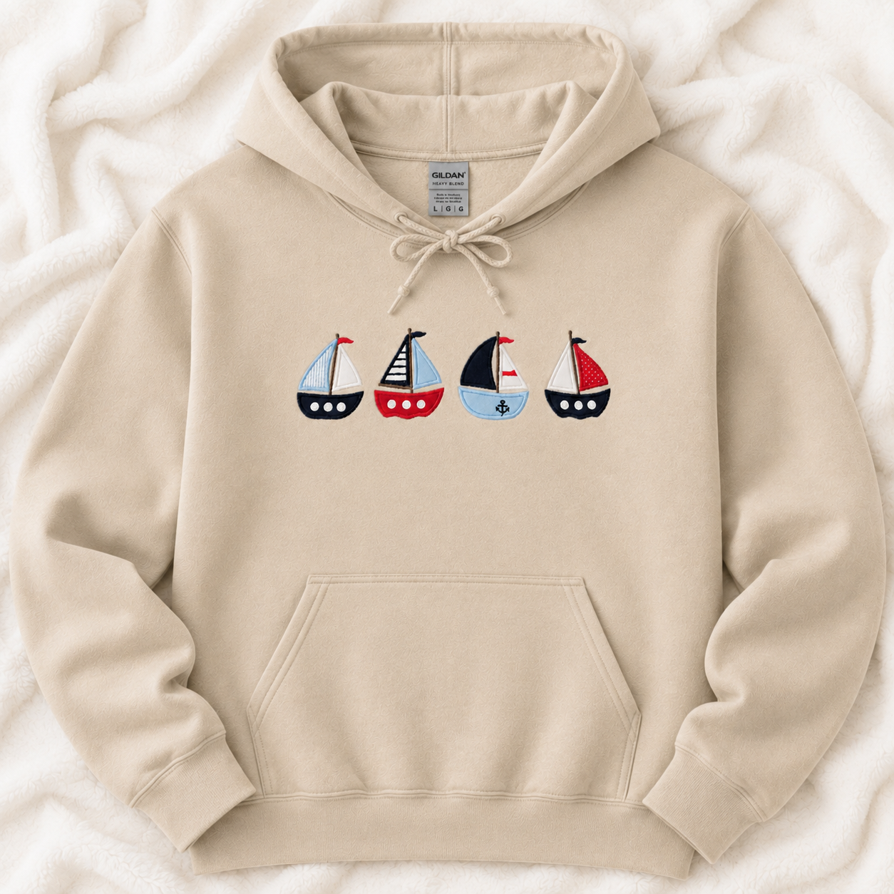 A sand pullover hoodie laid flat on a soft white textured blanket. The hoodie features a drawstring hood, a front kangaroo pocket, and a graphic of four sailboats across the chest in red, navy, and light blue.