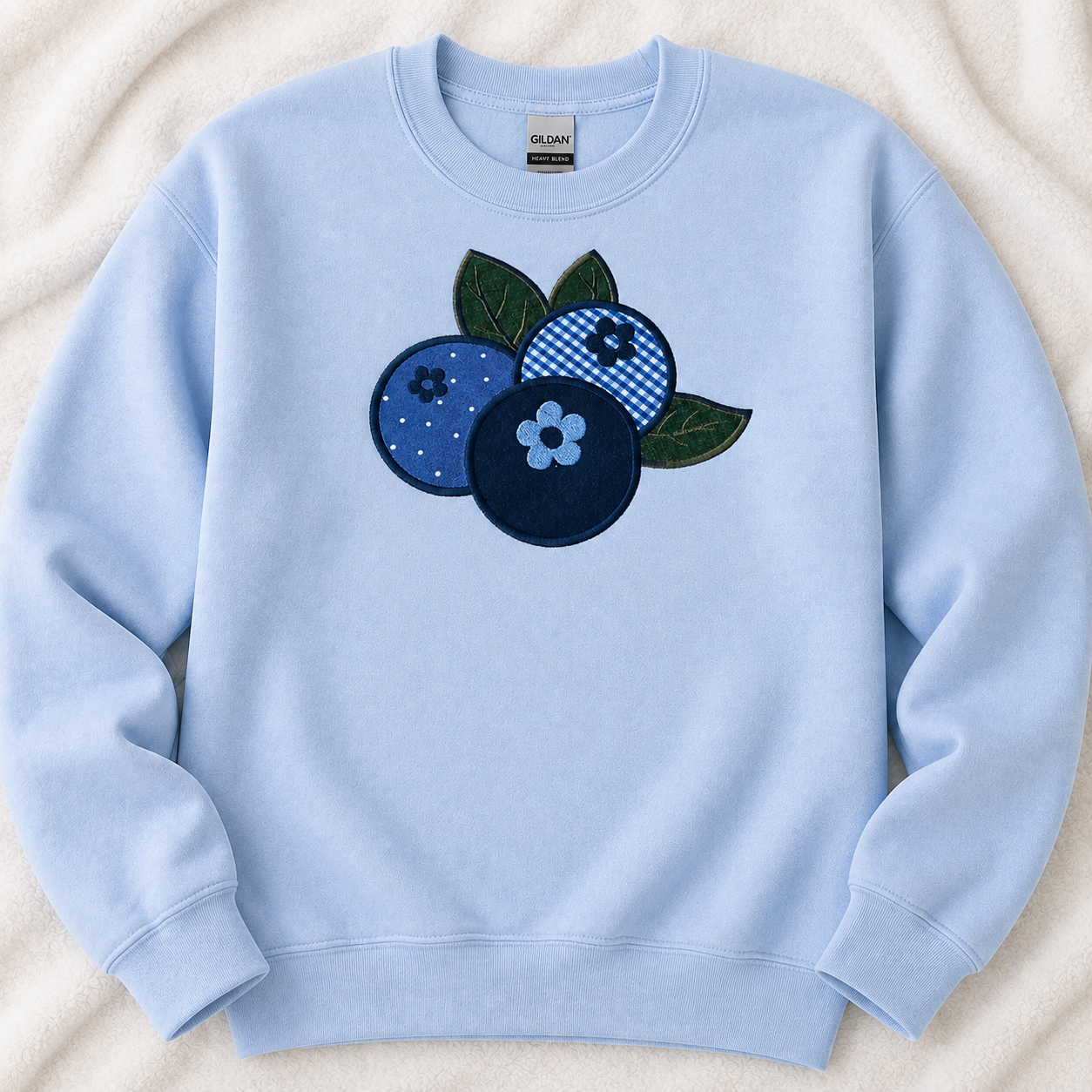 A light blue crewneck sweatshirt featuring a central blueberry applique with green leaves, laid flat on a textured cream fabric background.