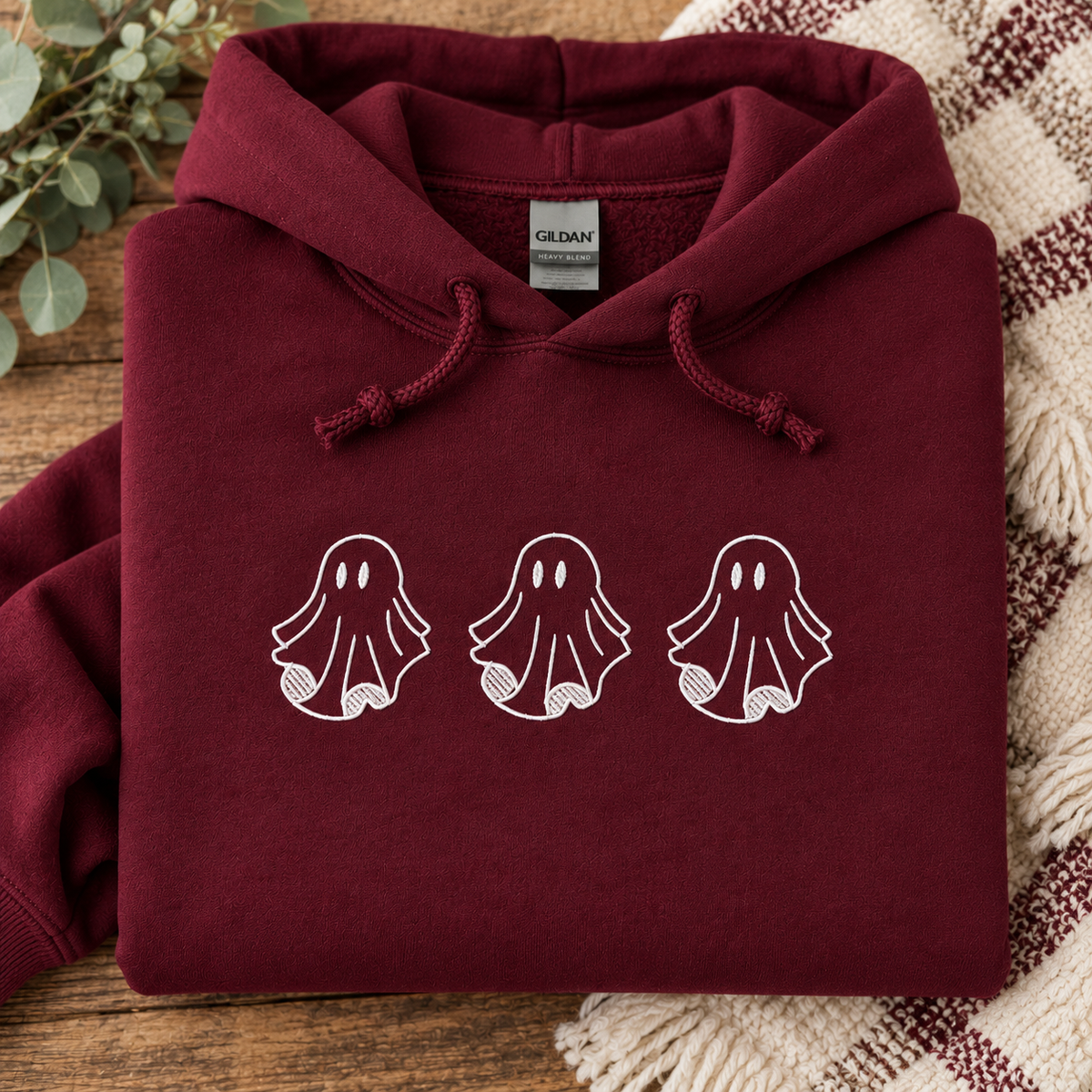 A folded maroon hoodie with three white ghost embroidered across the chest, displayed on a wooden surface with a knit blanket and eucalyptus leaves in the background. The inner neck label shows Gildan.