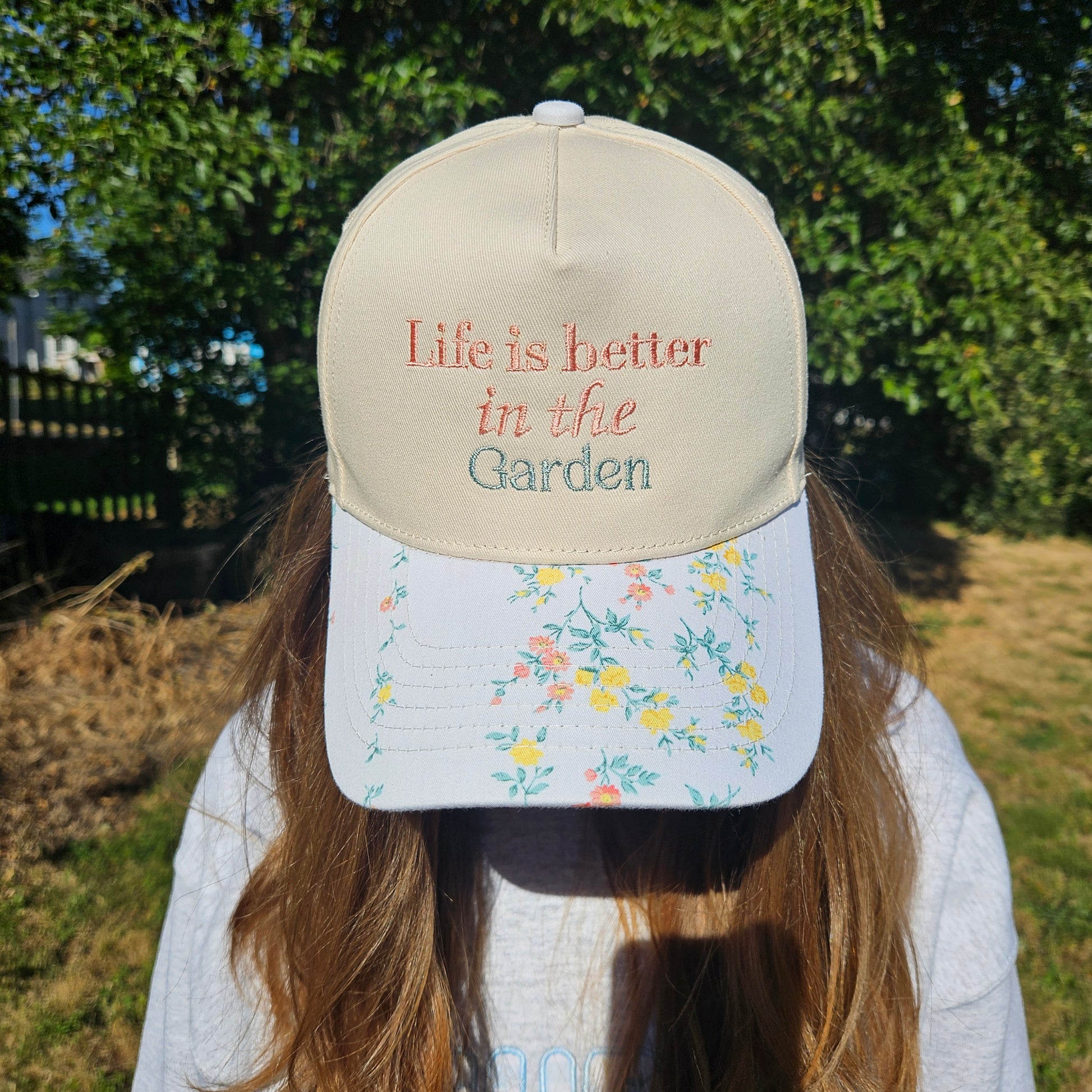 Person wearing a cap with 'Life is better in the garden' text and floral design outdoors.
