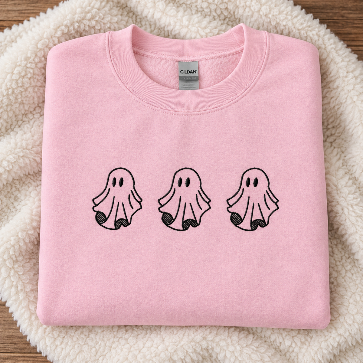 A folded light pink sweatshirt with three black ghost embroidered across the chest, displayed on a cream sherpa fabric background. The neck label shows the brand Gildan.
