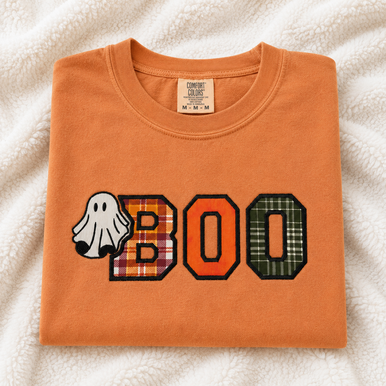 A yam Comfort Colors t-shirt folded neatly on a white textured fleece blanket. The front features a 'BOO' graphic with a white ghost and plaid patterned letters.