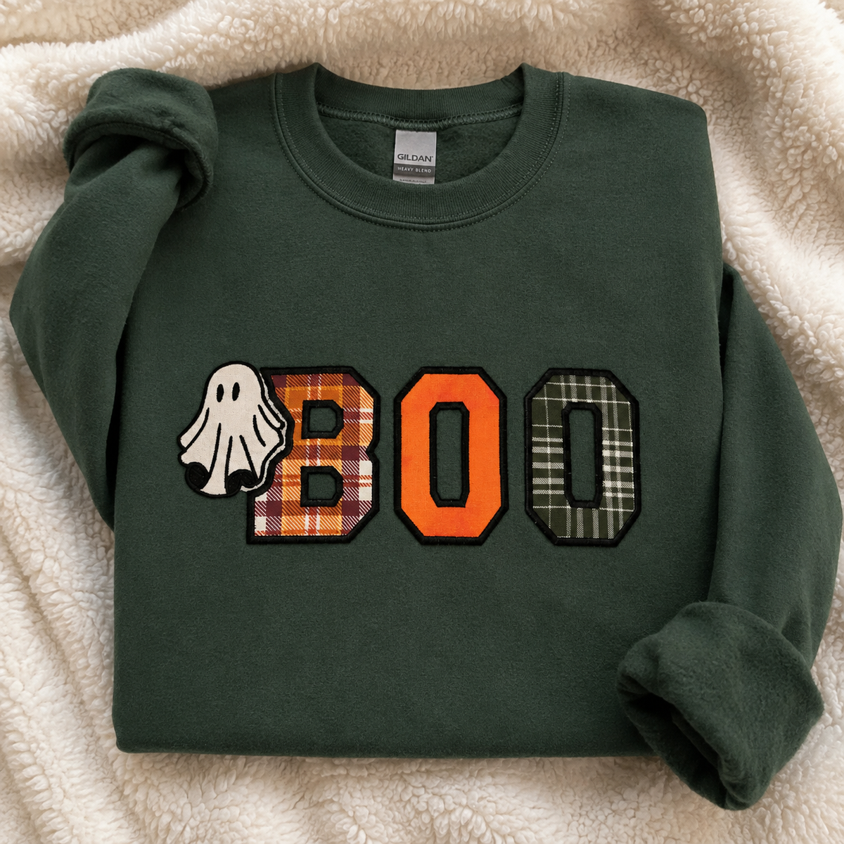 A forest green crewneck sweatshirt laid flat on a cream sherpa fleece background. The front features a 'BOO' graphic with a white ghost, a plaid patterned 'B', and an orange 'O', followed by a plaid patterned 'O'