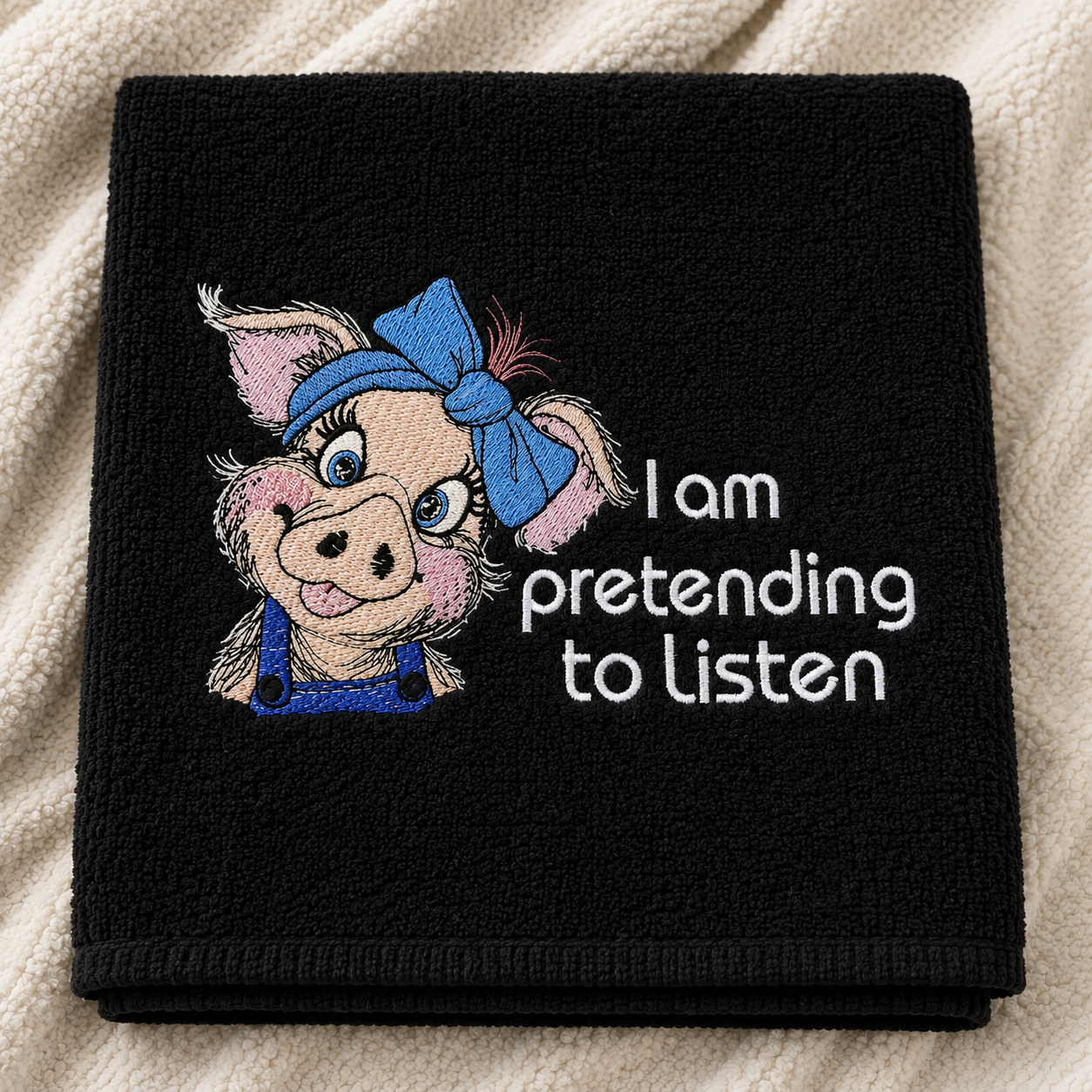 Black towel with embroidered cartoon pig and text 'I am pretending to listen' on a textured surface.