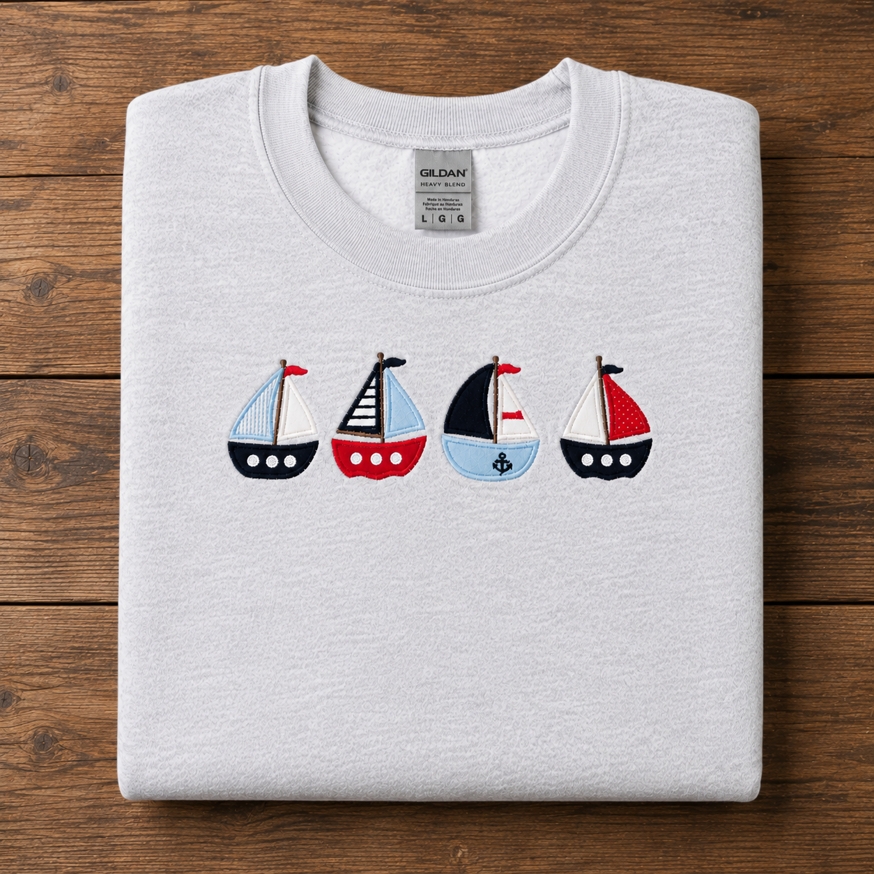 A folded ash Gildan sweatshirt with an applique of four colorful sailboats across the chest, displayed flat on a dark wood background.