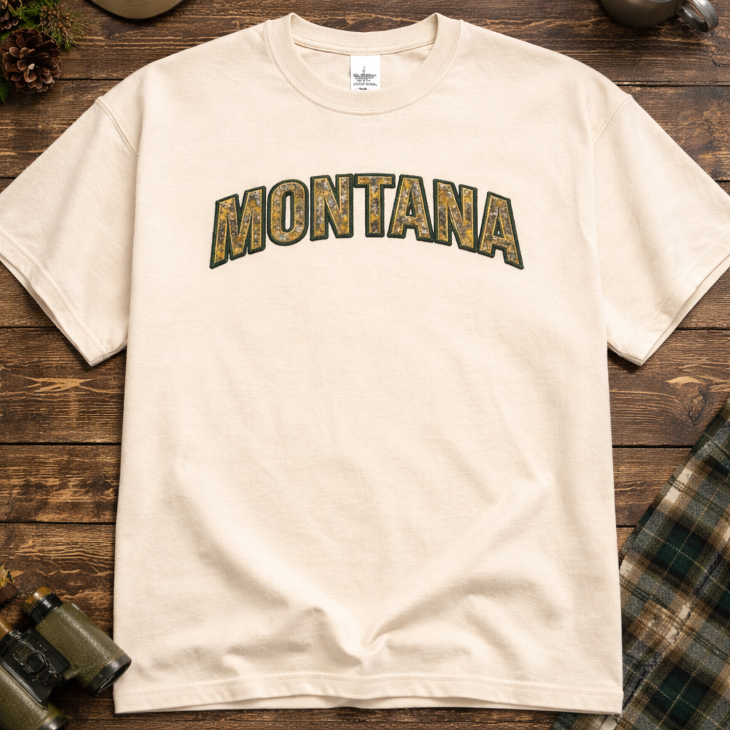Ivory t-shirt with applique 'MONTANA' embroidered on it, placed on a wooden surface with outdoor gear.