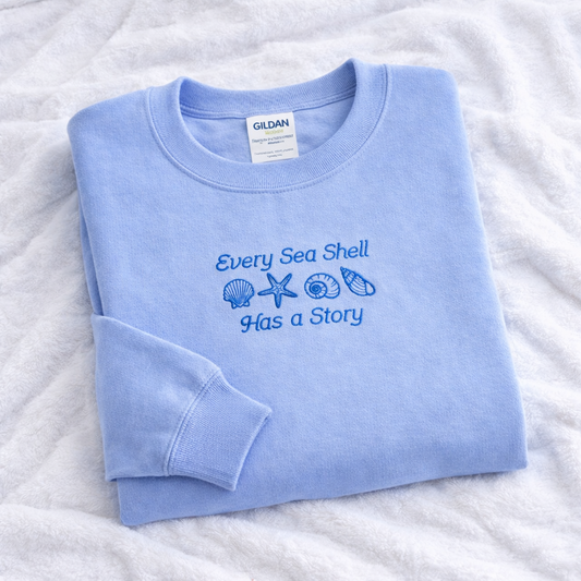 Folded light blue sweatshirt with 'Every Sea Shell Has a Story' text and design on a white textured background