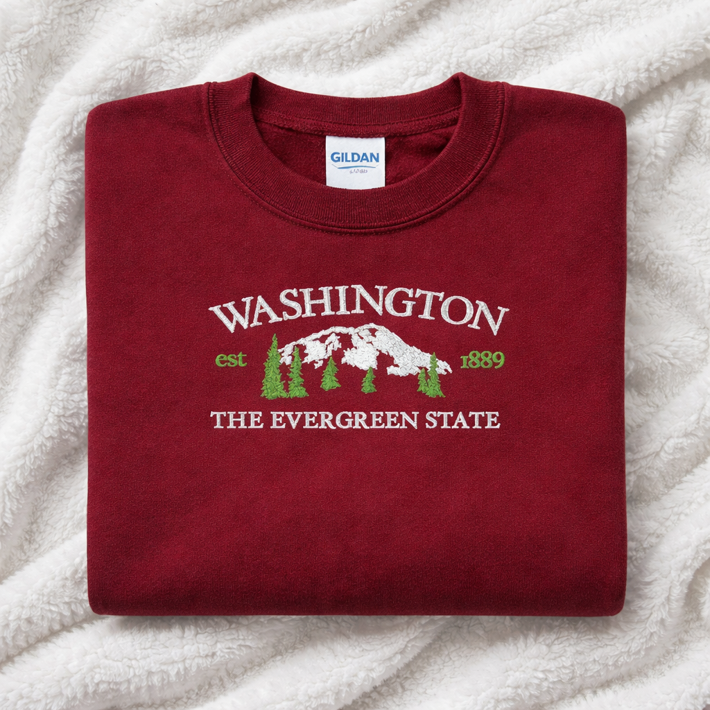 Folded maroon sweatshirt with 'Washington The Evergreen State' text and design on a white blanket background