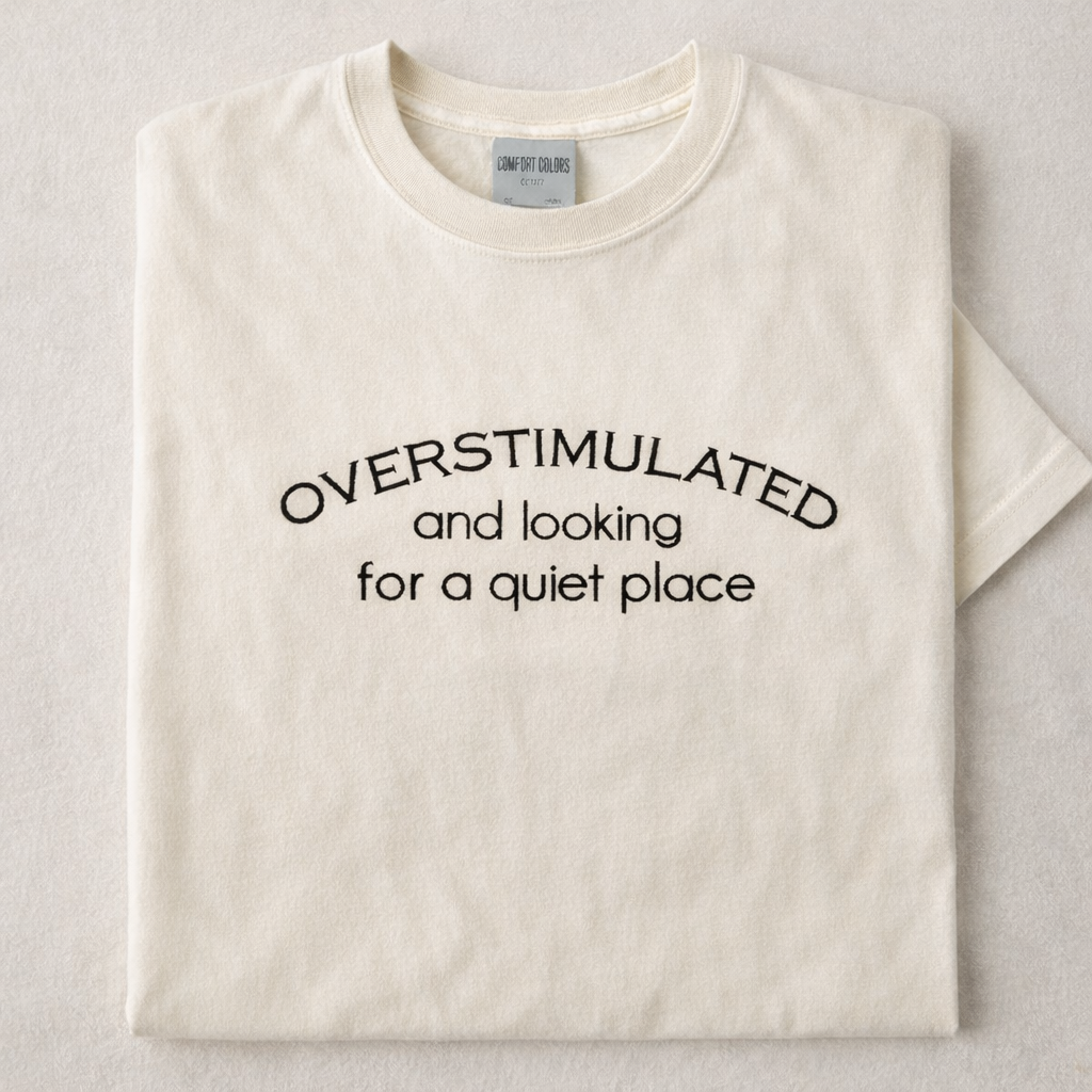 Ivory t-shirt with embroidered text 'OVERSTIMULATED and looking for a quiet place' on a white background