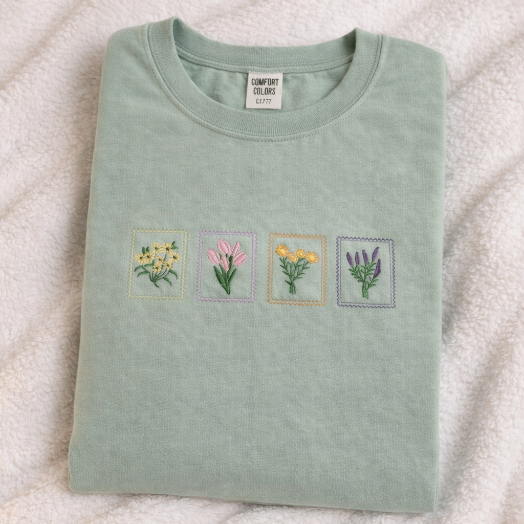 Folded sage t-shirt with embroidered floral designs on a textured white surface