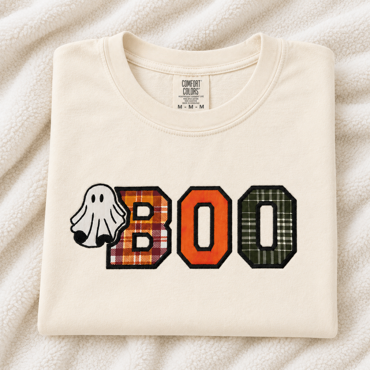 A folded ivory Comfort Colors t-shirt with a 'BOO' Halloween graphic on the front, featuring a ghost and plaid patterned letters, laid flat on a textured cream fleece background.