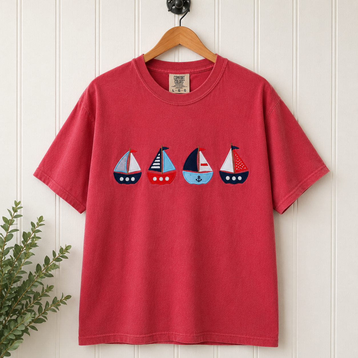 Crimson t-shirt with sailboat design on a wooden hanger against a white wall.