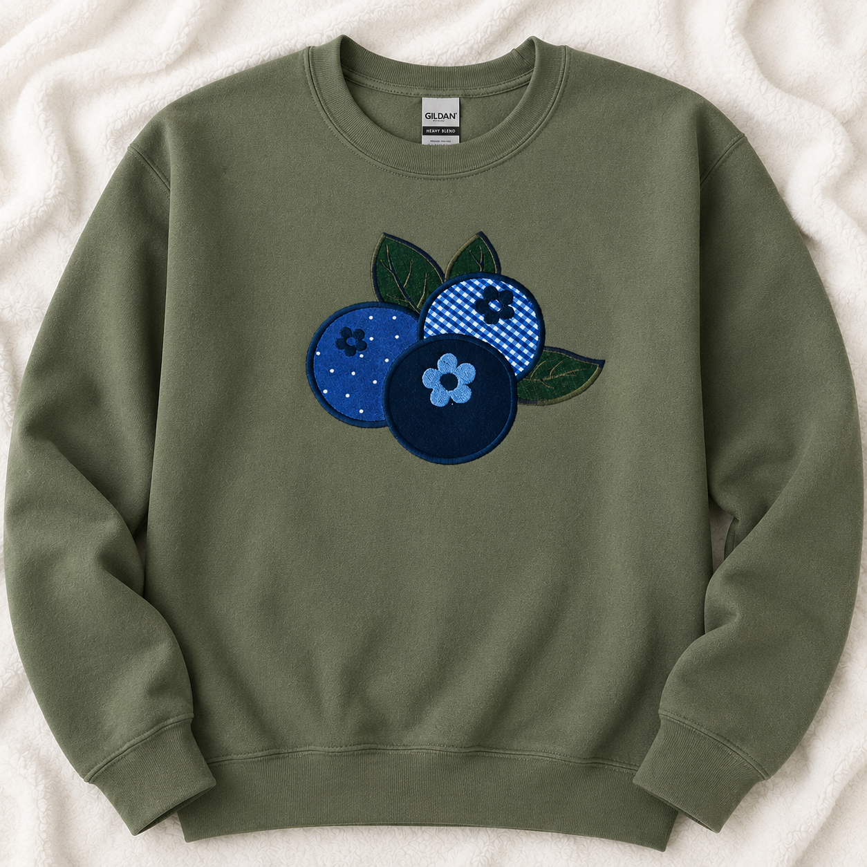 An militry green crewneck sweatshirt featuring an applique design of three blueberries in various patterns (solid blue, checkered blue-and-white, and navy with a light blue flower) on the chest, laid flat on a white textured blanket. The brand label 'GILDAN' is visible inside the collar.