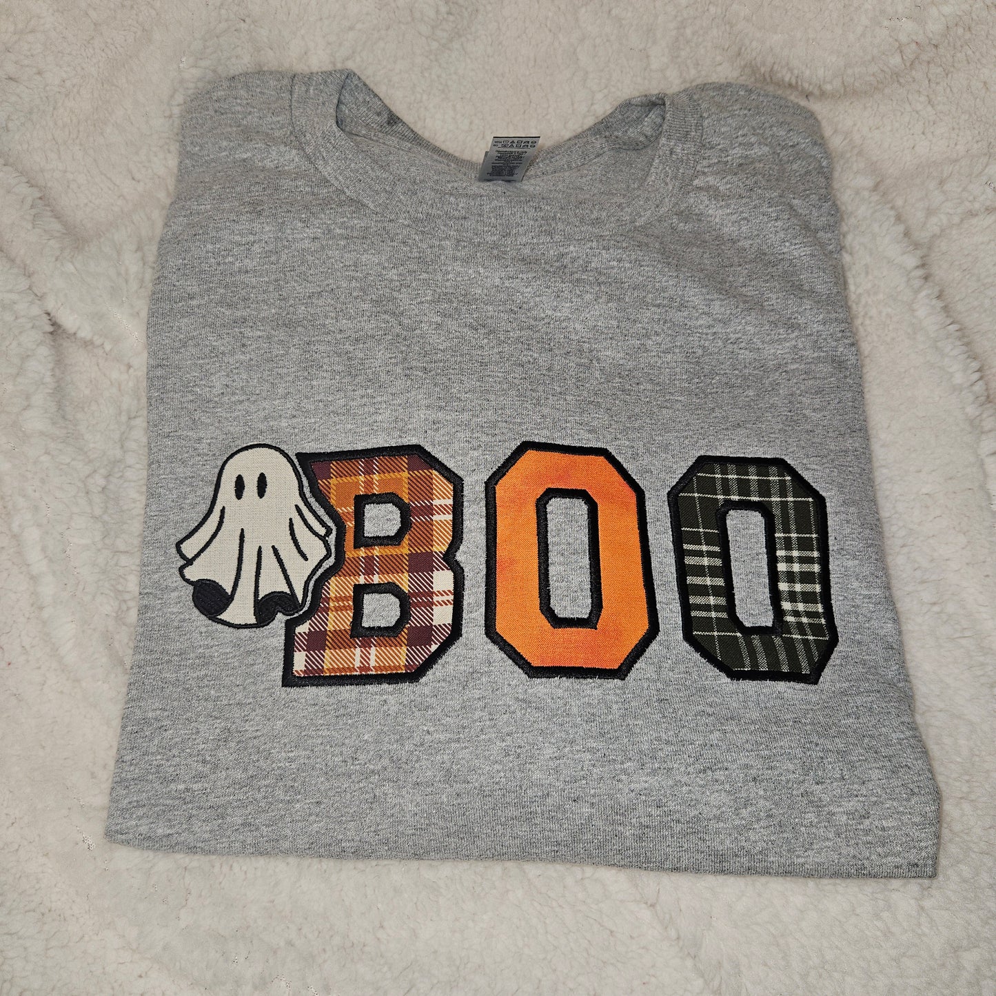A folded heather gray t-shirt featuring a 'BOO' graphic with a ghost, orange plaid letter 'B', solid orange letter 'O', and black-and-white plaid letter 'O' on a white faux fur background.