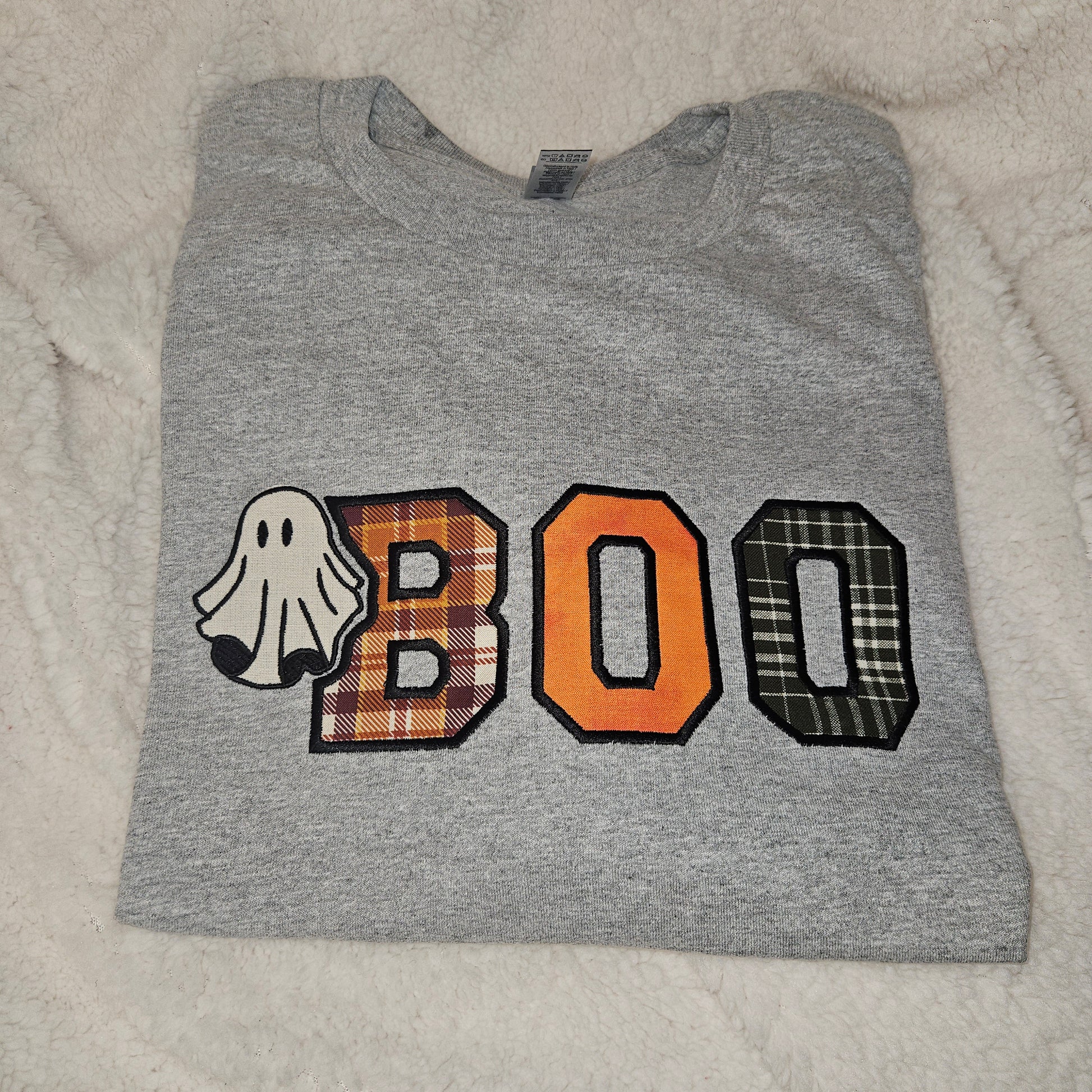 A folded heather gray t-shirt featuring a 'BOO' graphic with a ghost, orange plaid letter 'B', solid orange letter 'O', and black-and-white plaid letter 'O' on a white faux fur background.