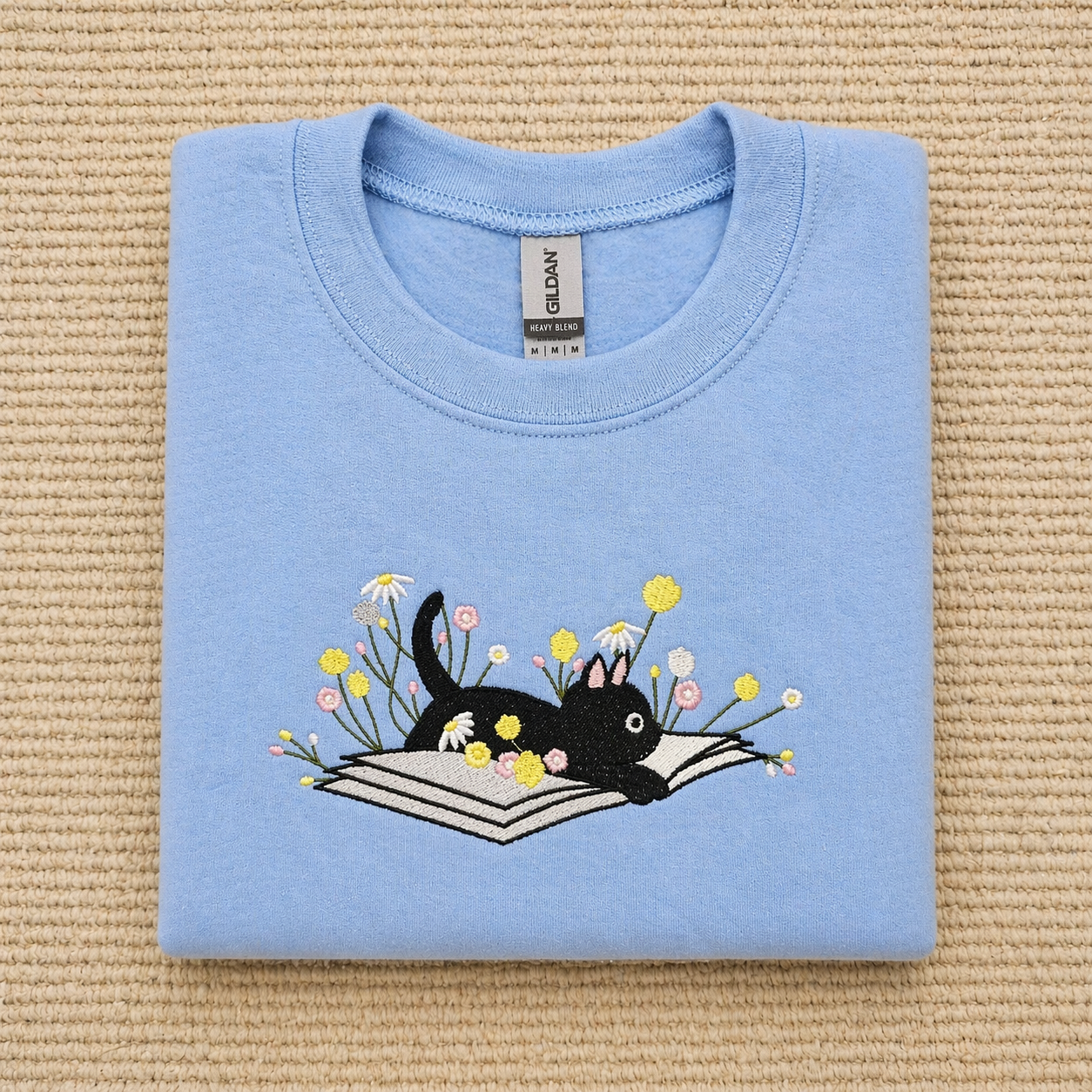 Folded carolina blue sweatshirt with a black cat and flowers design on a textured beige surface
